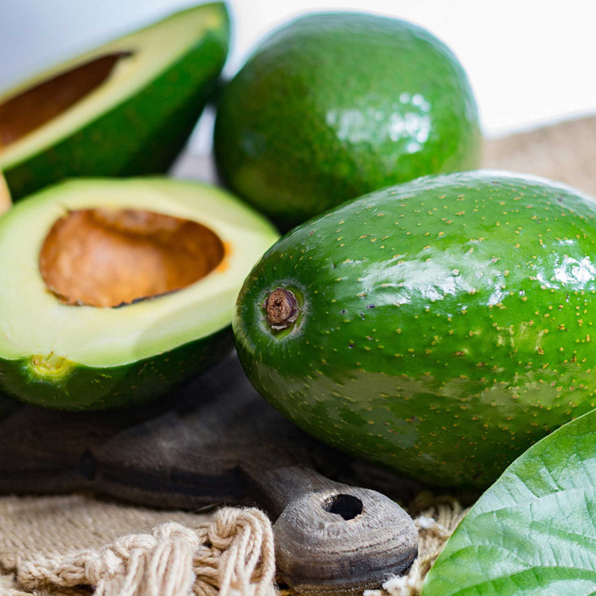 Fresh green avocados on a rustic wooden board with burlap and a leaf, including one halved avocado showing the brown pit.