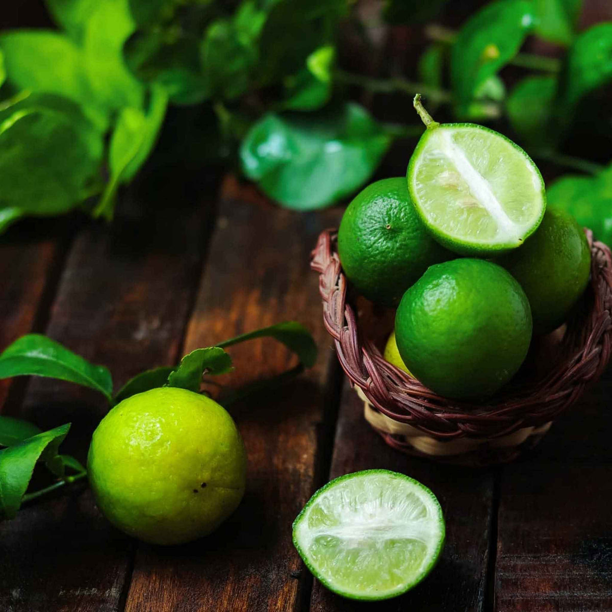 Fresh green key limes, including whole and halved pieces, arranged in a small woven basket and on a dark wooden surface with green leaves in the background.