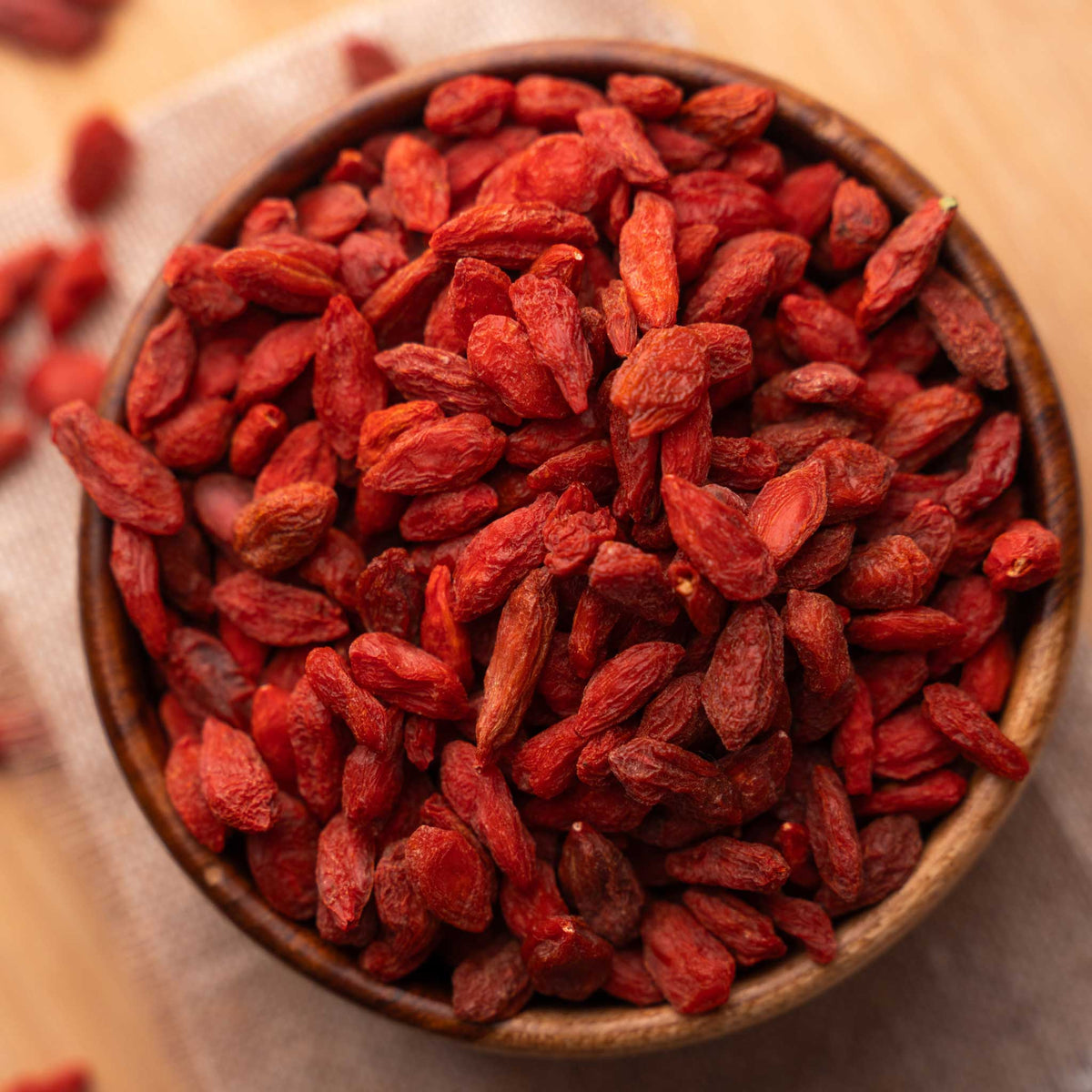 A wooden bowl filled with dried red goji berries on a light fabric surface with a few scattered in the background.