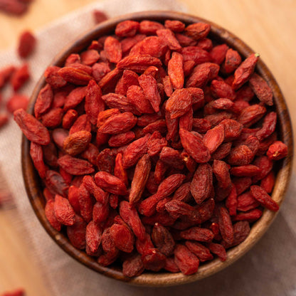 A wooden bowl filled with dried red goji berries on a light fabric surface with a few scattered in the background.