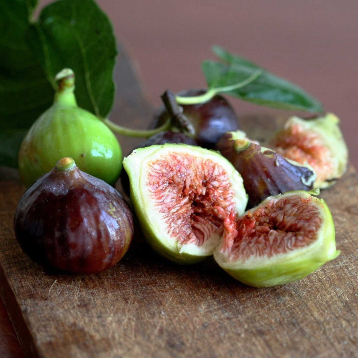 Fresh little miss figgy figs on a rustic wooden cutting board, including whole dark purple figs and two halves cut open to show pinkish-red seeds and pale green flesh, with green leaves in the background.