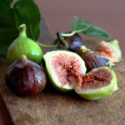 Fresh little miss figgy figs on a rustic wooden cutting board, including whole dark purple figs and two halves cut open to show pinkish-red seeds and pale green flesh, with green leaves in the background.