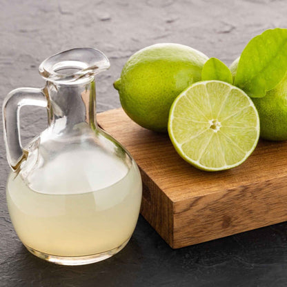 Clear glass container with a liquid next to whole and sliced persian bearss limes on a wooden board.